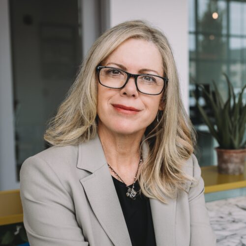 A person with long blonde hair and glasses wearing a beige blazer sits indoors. A plant is visible in the background.