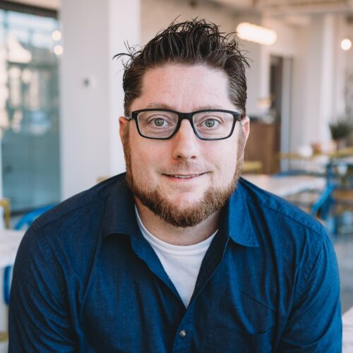 Man with glasses and a beard wearing a blue shirt sits indoors, looking at the camera.