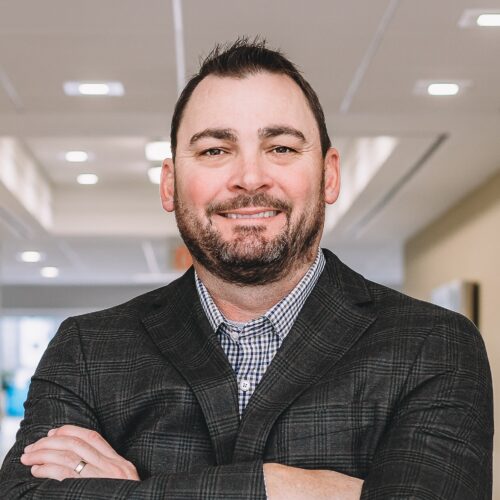 A man with short hair and a beard, wearing a plaid jacket and checked shirt, smiles confidently with arms crossed in a well-lit hallway.