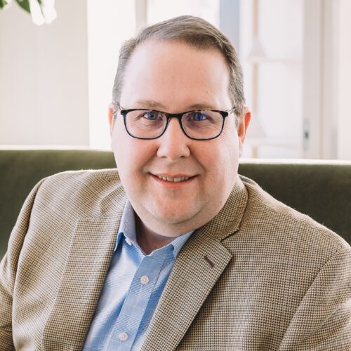 A man in glasses and a beige blazer smiles at the camera in a well-lit room.