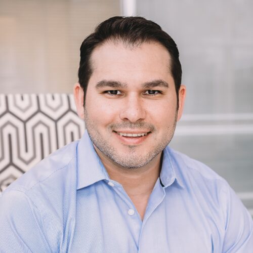 Man in a light blue shirt smiles while sitting indoors with a patterned chair in the background.