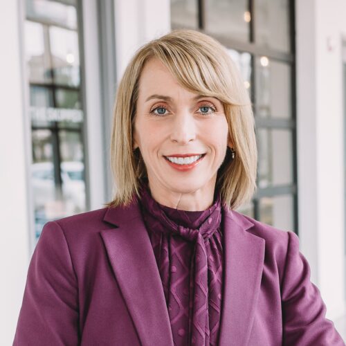 Woman with short blonde hair in a purple blazer and blouse, smiling, stands in a modern interior with large windows.