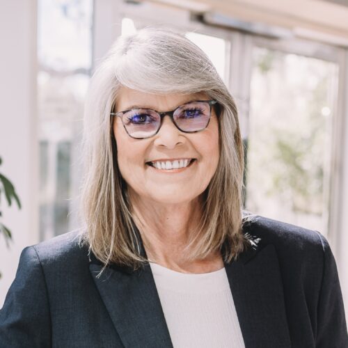 Smiling woman with gray hair and glasses wearing a navy blazer and white top, standing indoors with a bright, blurred background.