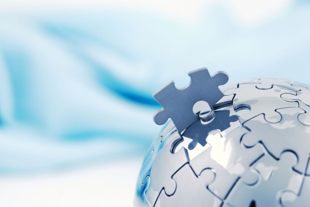 A metallic globe with puzzle pieces has one piece being inserted at the top. The background is a blurred light blue.