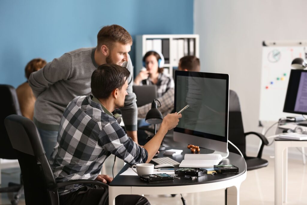 Two men in a modern office reviewing content on a computer monitor. Three additional people work in the background at desks with laptops and large screens.