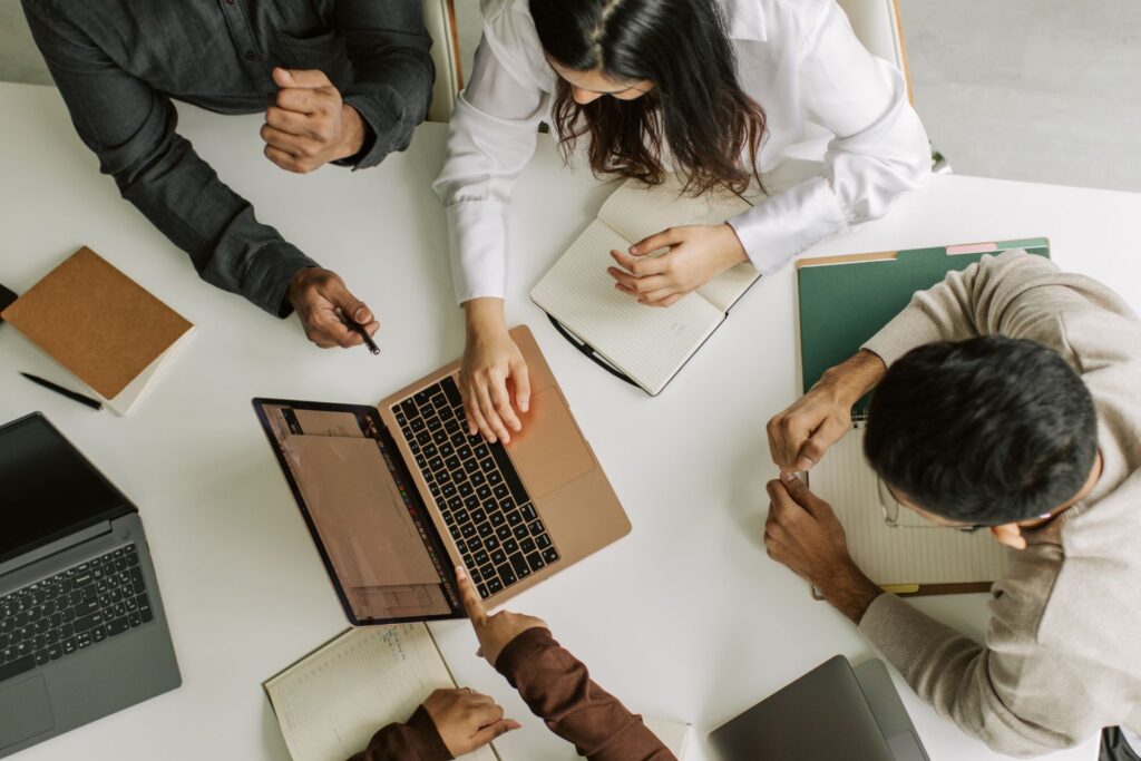 Four people collaborating around a table with laptops, notebooks, and coffee mugs, viewed from above.