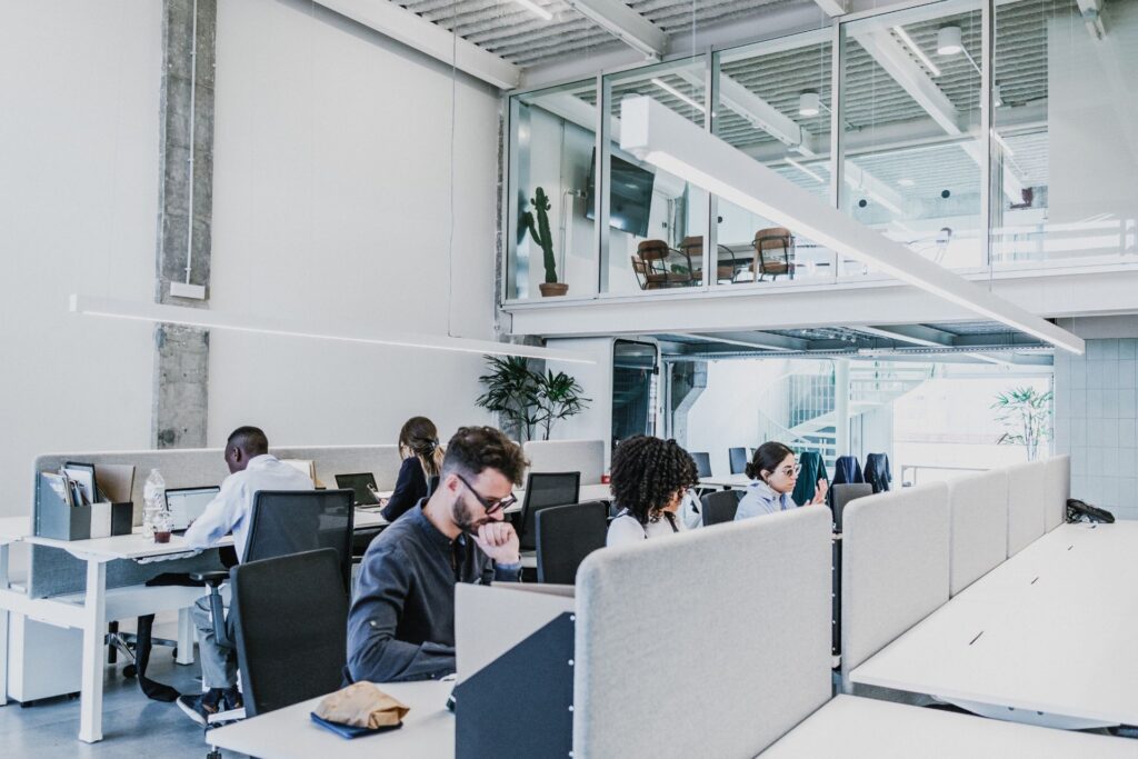 Open-plan office with people working at desks; a mezzanine level visible above with additional seating and plants.