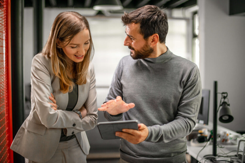 A man shows a woman something on a tablet while they stand in an office and have a discussion.