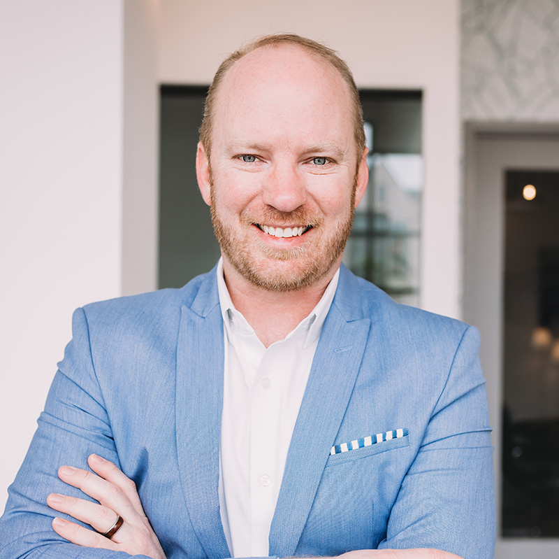 A man with a beard and short hair, wearing a light blue suit and white shirt, stands smiling with his arms crossed in a modern indoor setting, giving off an approachable aura perfect for an about page.