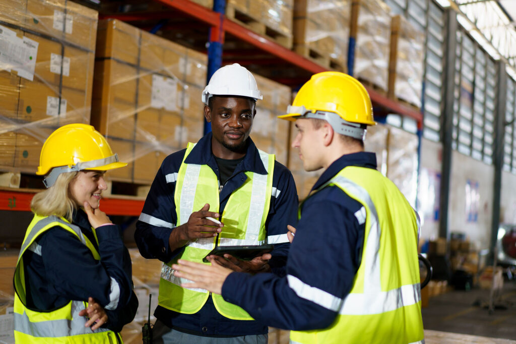 Three workers in safety gear and helmets stand talking in a warehouse, surrounded by shelves of boxed goods. One holds a tablet while another gestures during the discussion.