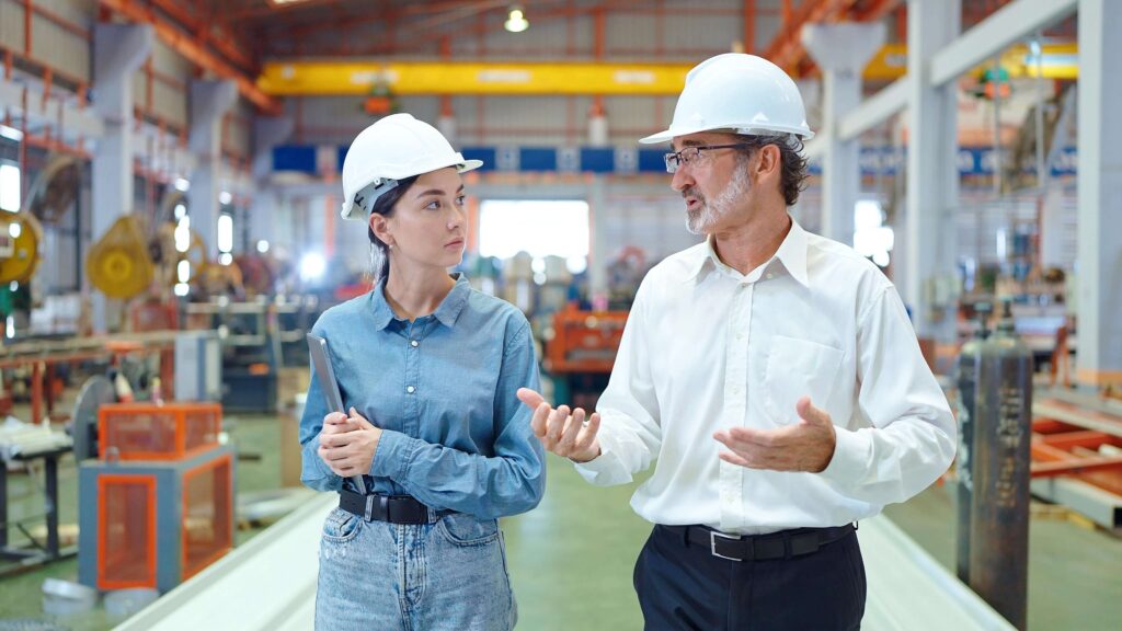 Two people wearing safety helmets and business attire are having a discussion inside a large industrial facility or factory.