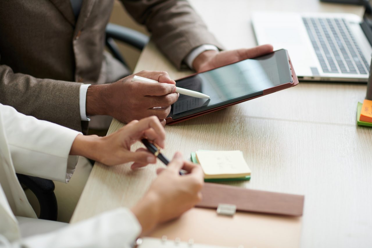 Two people at a desk review information on a tablet, with notebooks, sticky notes, and a laptop nearby.