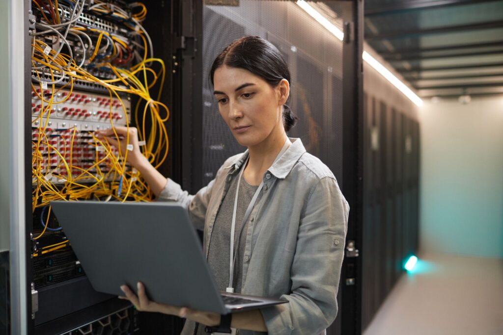 A person holding a laptop works on a server rack with numerous cables in a cloud data center.