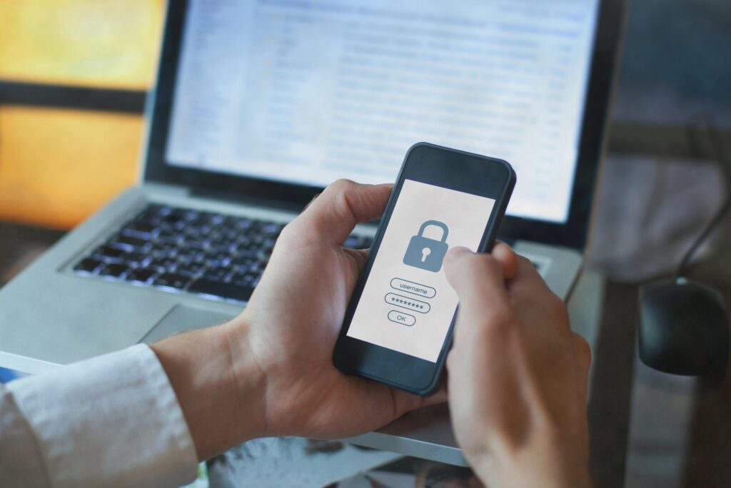 Cybersecurity: Person holding a smartphone showing a padlock icon and password login screen, with a laptop computer in the background.