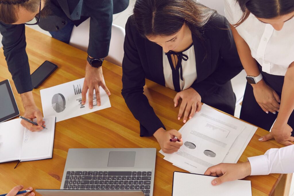 Four people in business attire work together at a wooden table with charts, papers, notebooks, a laptop, and smartphones.