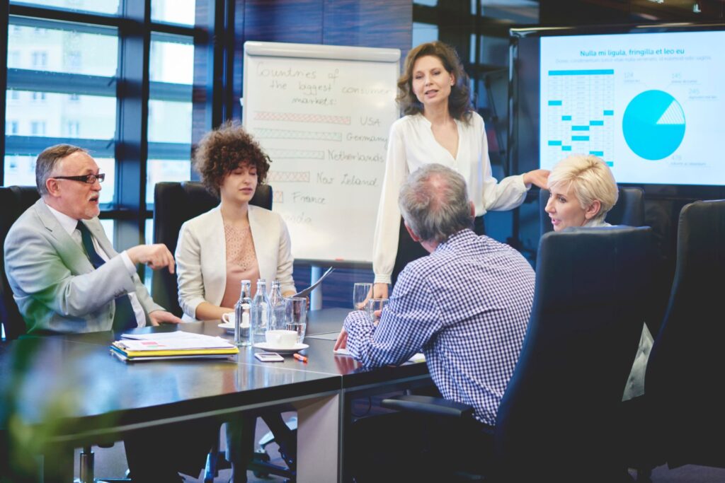 Five people in business attire have a meeting in a conference room with charts on a screen and flipchart, discussing documents and graphs.