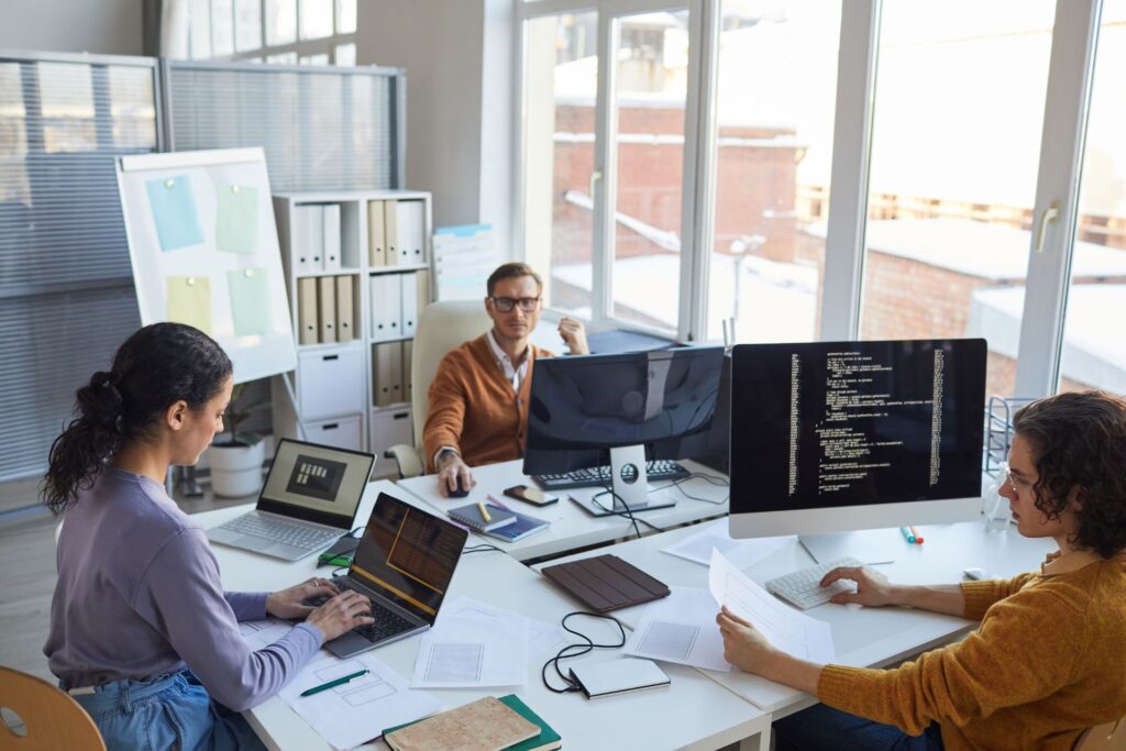 Three people work at desks with computers and documents in a modern office, focusing on IT cost optimization, with large windows, shelves, and folders visible in the background.