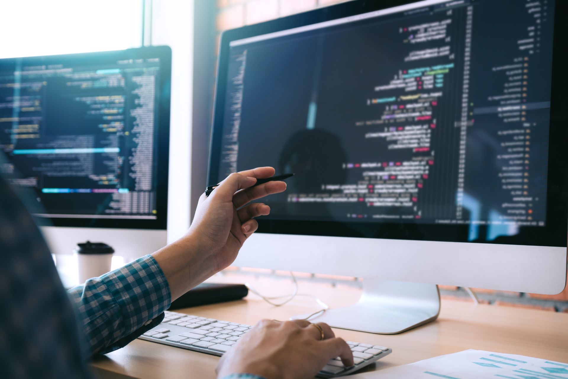 A person in a plaid shirt points at lines of code on a large computer monitor, discussing IT cost optimization strategies, while another screen displaying code sits in the background on a desk.