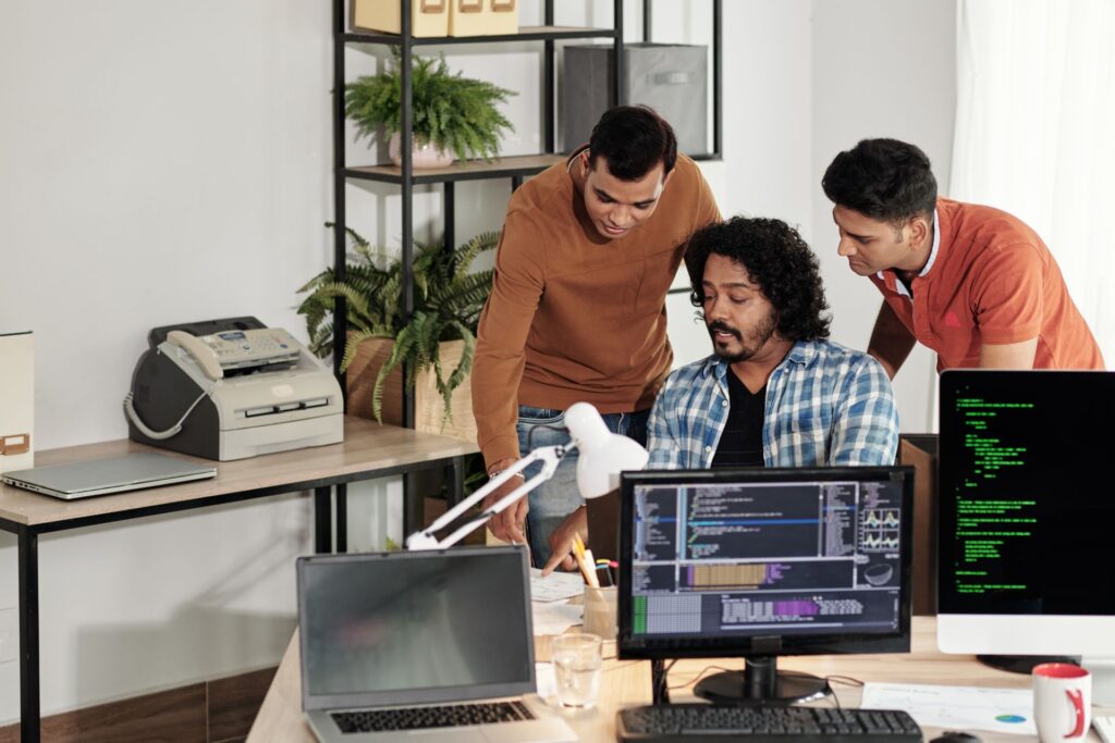 Three men in a modern office collaborate at a desk with multiple monitors displaying code and IT cost optimization strategies; plants and office equipment are visible in the background.