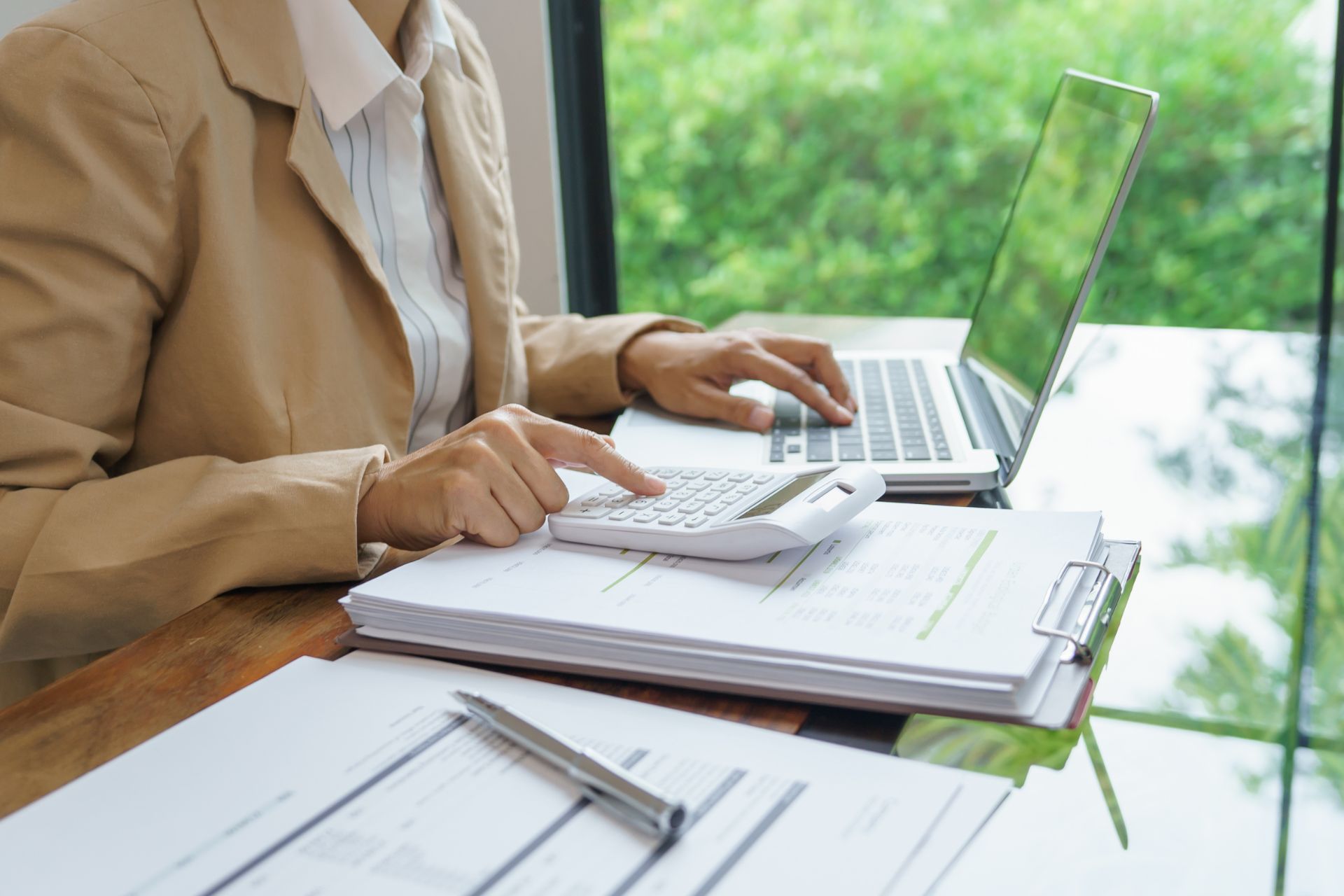 Person in a beige blazer using a laptop and calculator at a desk with documents, clipboard, and pen, focused on IT Cost Optimization, with greenery visible through a window in the background.