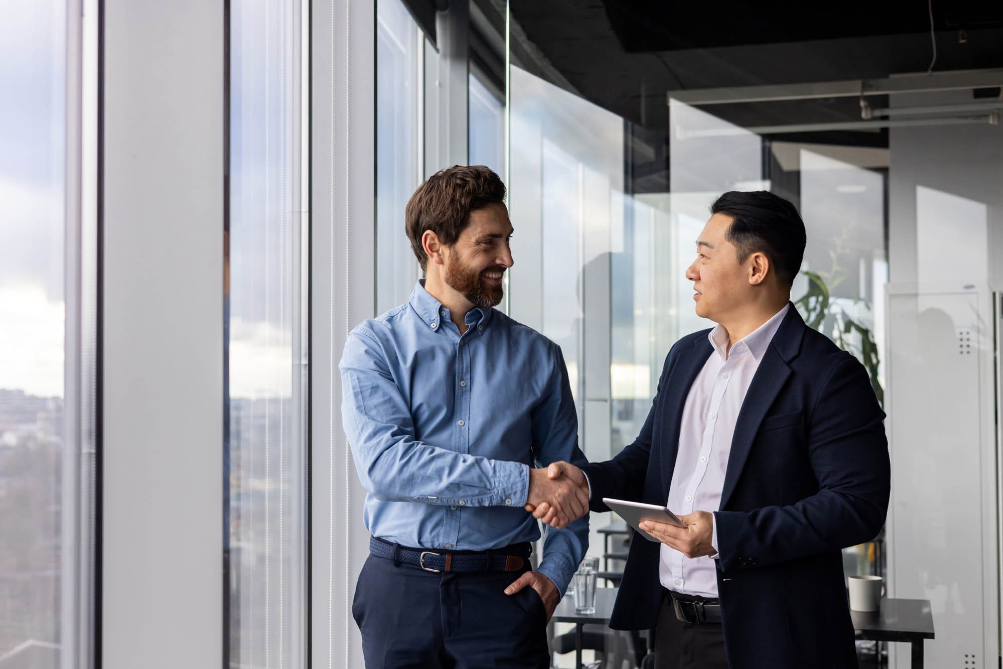 Business Partners: Two men in business attire shake hands in a modern office with large windows. One holds a tablet.