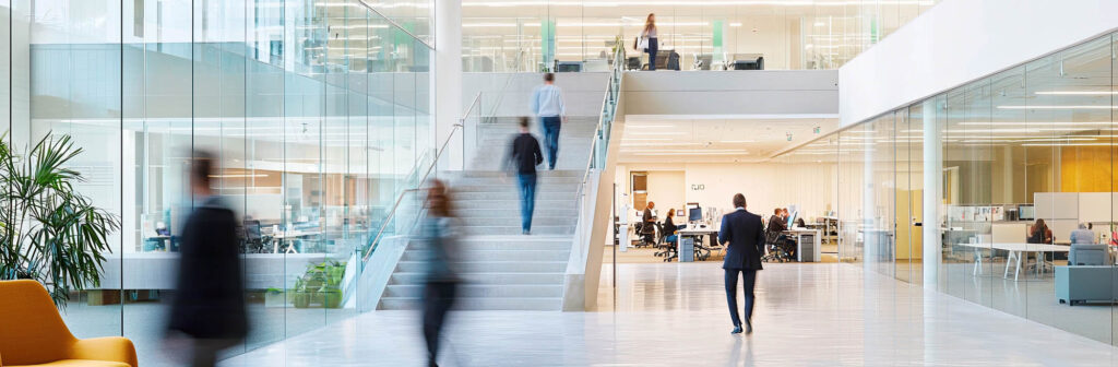 A modern office interior with people walking and working. Large glass walls, open space, and a staircase leading to an upper level are visible.