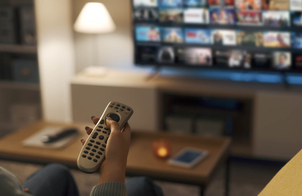 Person holding a remote control directed at a television displaying a streaming service interface, with a lit table lamp and a tablet on a wooden coffee table in the background.