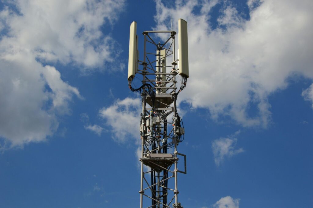 A cellphone tower belonging to a Network Service Provider stands tall, its antennas and cables reaching upward against a blue sky with scattered clouds.