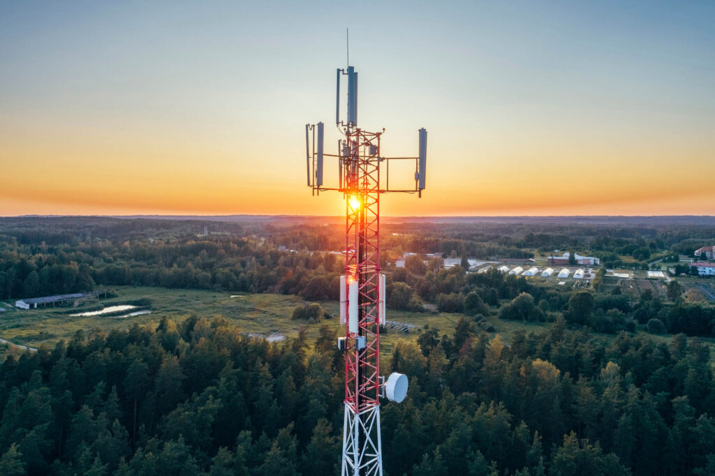 A telecommunications tower stands tall amidst a forested landscape at sunset, with the sun partly visible behind the structure.