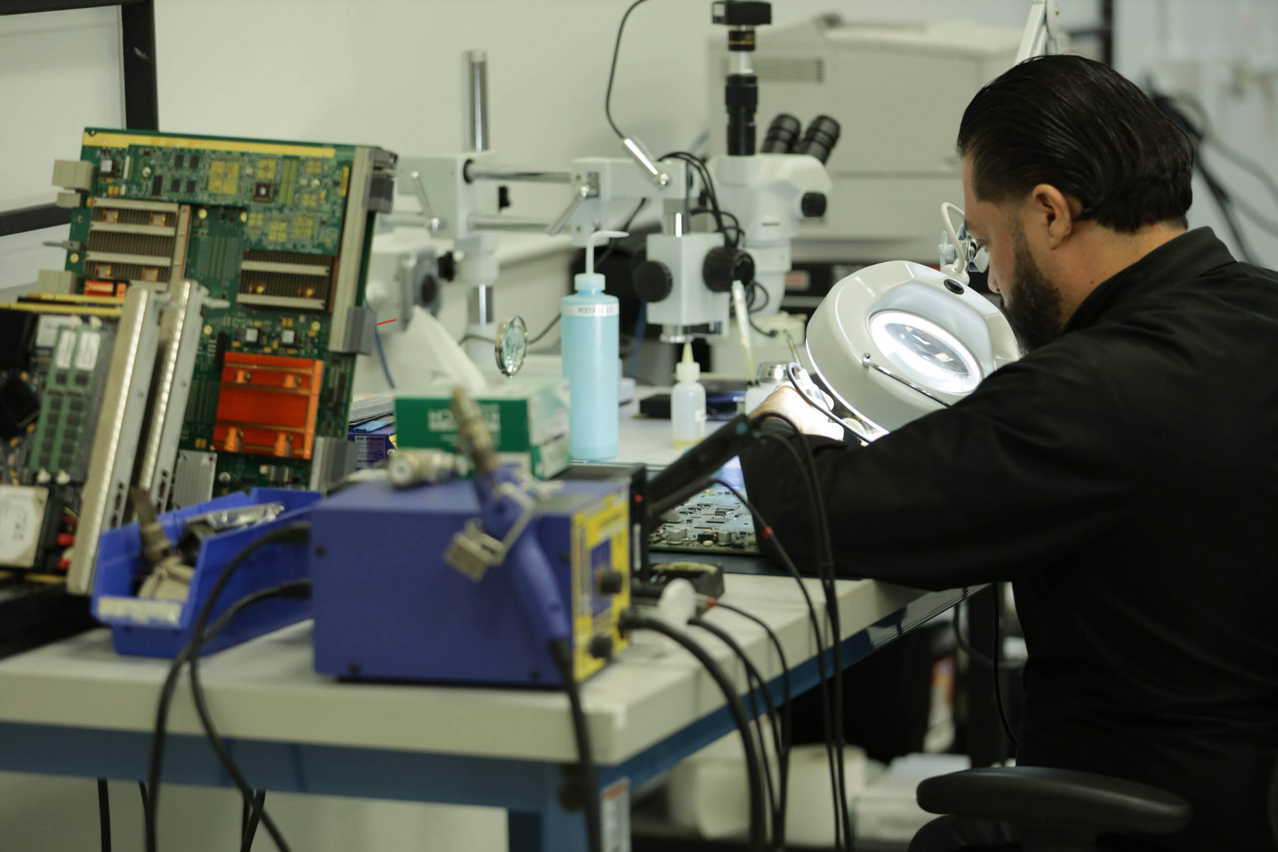 A technician, specializing in decommissioning services, works meticulously on electronic circuit boards at a workstation equipped with precision tools and a magnifying lamp.