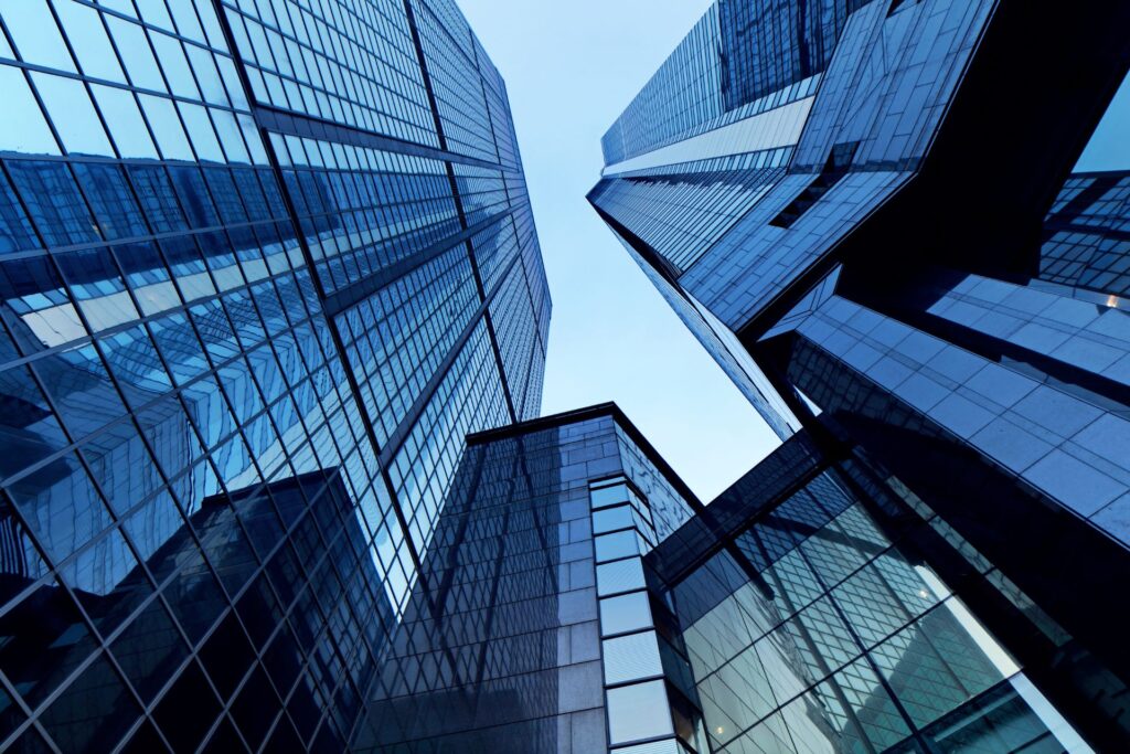 Tall modern glass skyscrapers reflect the sky and neighboring buildings, viewed from a low angle against a clear blue sky.