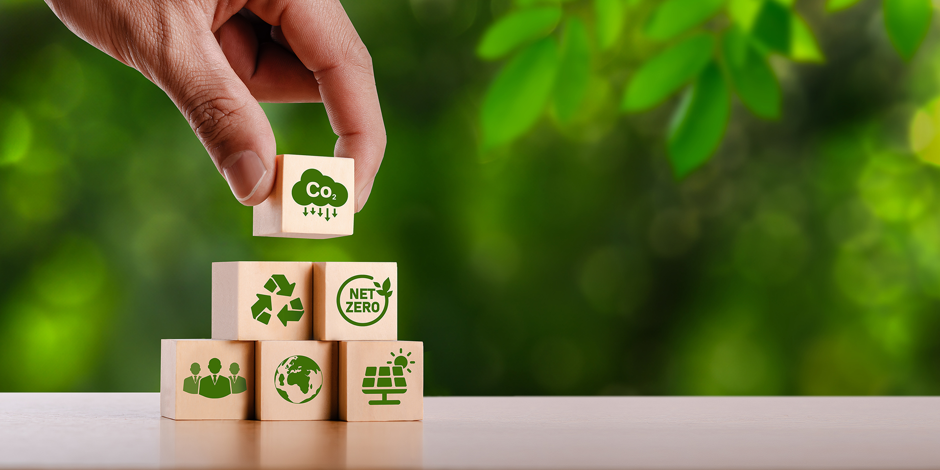 A hand places a wooden block with a CO2 icon on top of stacked blocks showing recycling, net zero, solar power, earth, and people symbols, with a green leafy background.