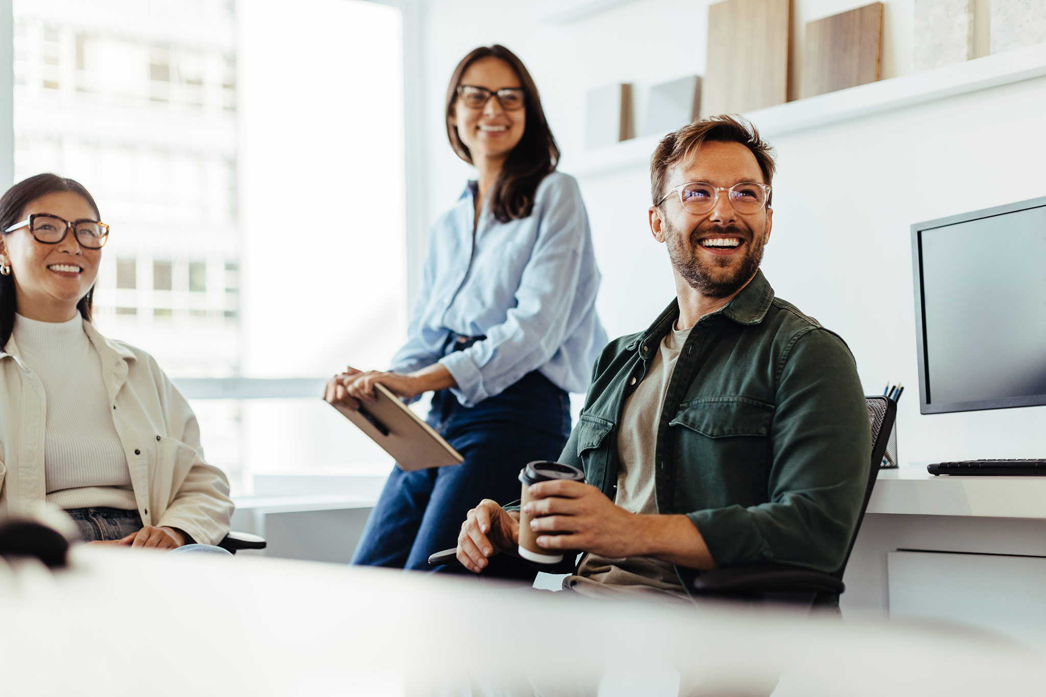 Three people in casual clothing sit and smile in a bright, modern office. One person holds a tablet, another holds a coffee cup. There are shelves and a computer monitor in the background.