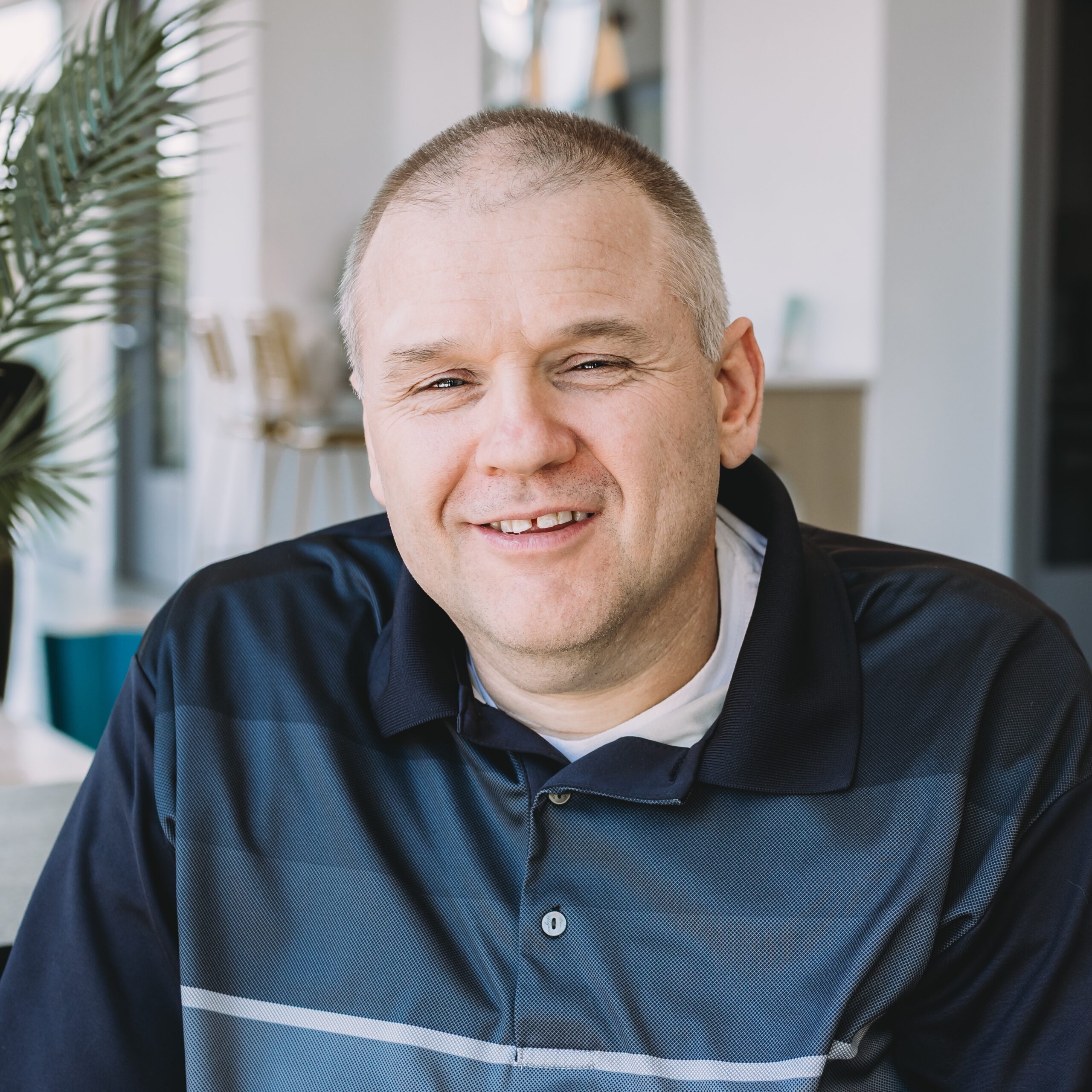 A middle-aged man with short hair and a slight smile sits indoors near a plant, wearing a dark polo shirt, appearing relaxed and about to engage in conversation.
