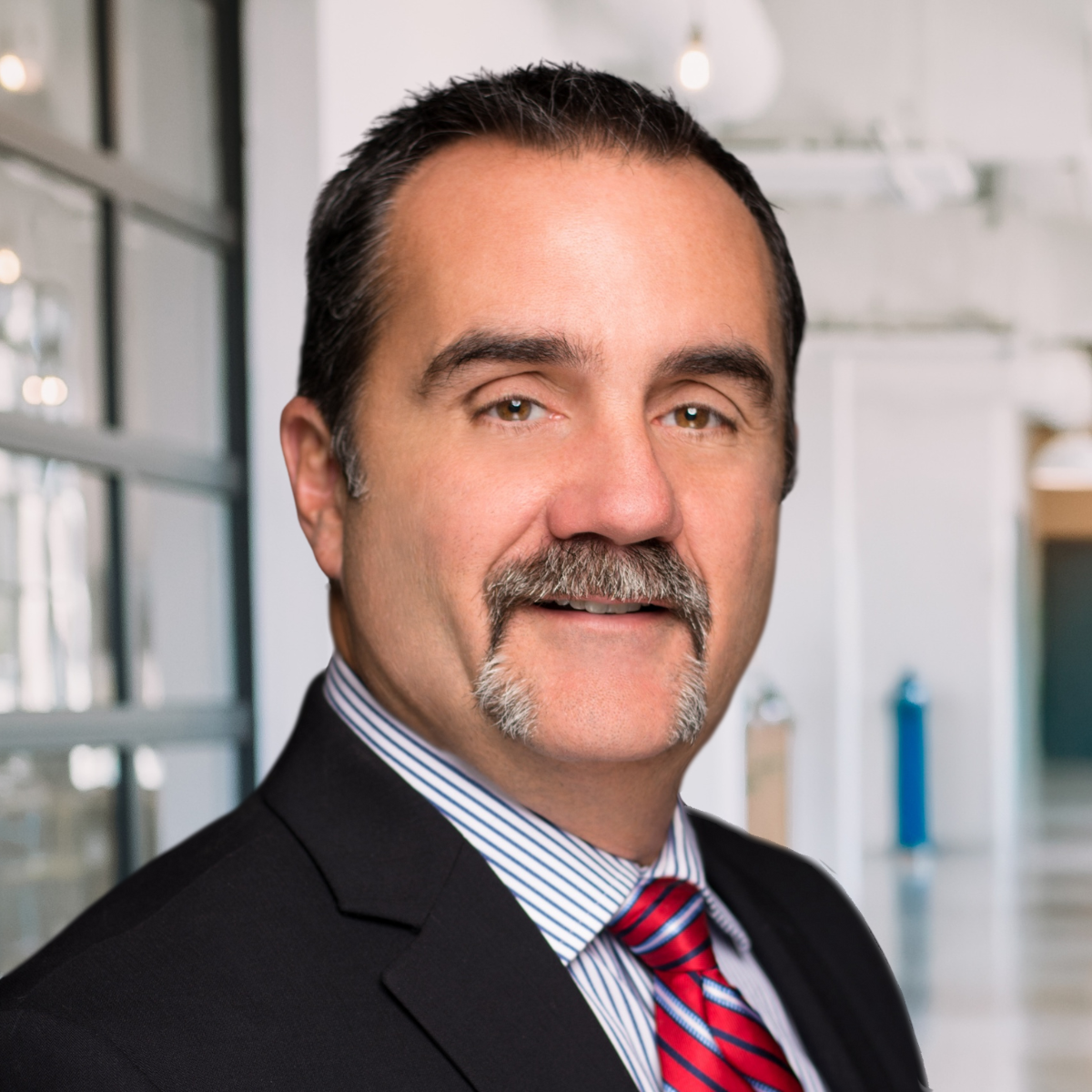 A man in a suit and red striped tie stands in a modern office hallway, looking at the camera with a neutral expression, conveying a professional look perfect for an "about" page.