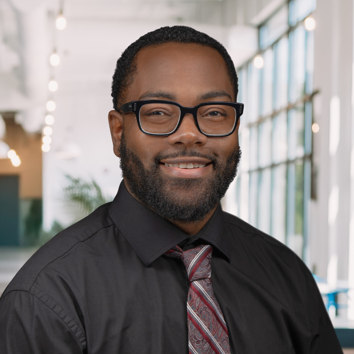 A man with short black hair, a beard, and glasses wearing a black shirt and patterned tie, smiling in a brightly lit modern office setting, is pictured in this photo about professional style.